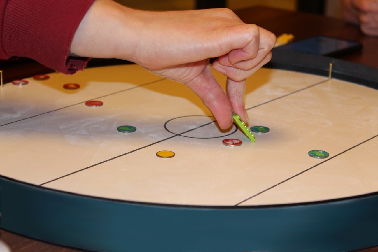 Nahaufnahme eines Spielbretts, auf dem eine Person mit einer Zange bunte Spielsteine bewegt. Es handelt sich um ein therapeutisches oder motorikförderndes Spiel – die Szene deutet auf eine Aktivierung oder Beschäftigung in der Pflege hin.